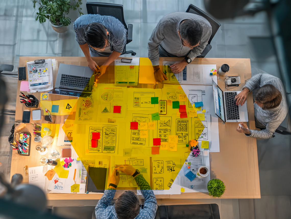 a top-down view of a desk as a team works on a website redesign. They sit around the space, looking at printed documents laid out on the table. Representing content, design, development, UX, and SEO teams working together.